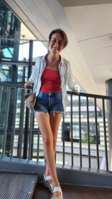 Junge Frau in rotem Top und Shorts, steht lächelnd an einem Geländer.