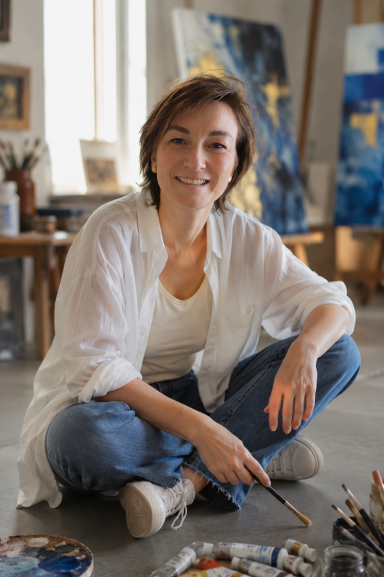 Junge Frau mit Pinsel in der Hand sitzt auf dem Boden in einem Atelier.